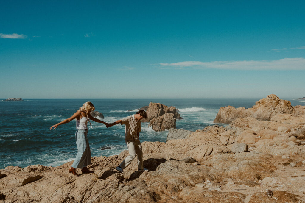 young couple holding hands walking along cliff by the ocean